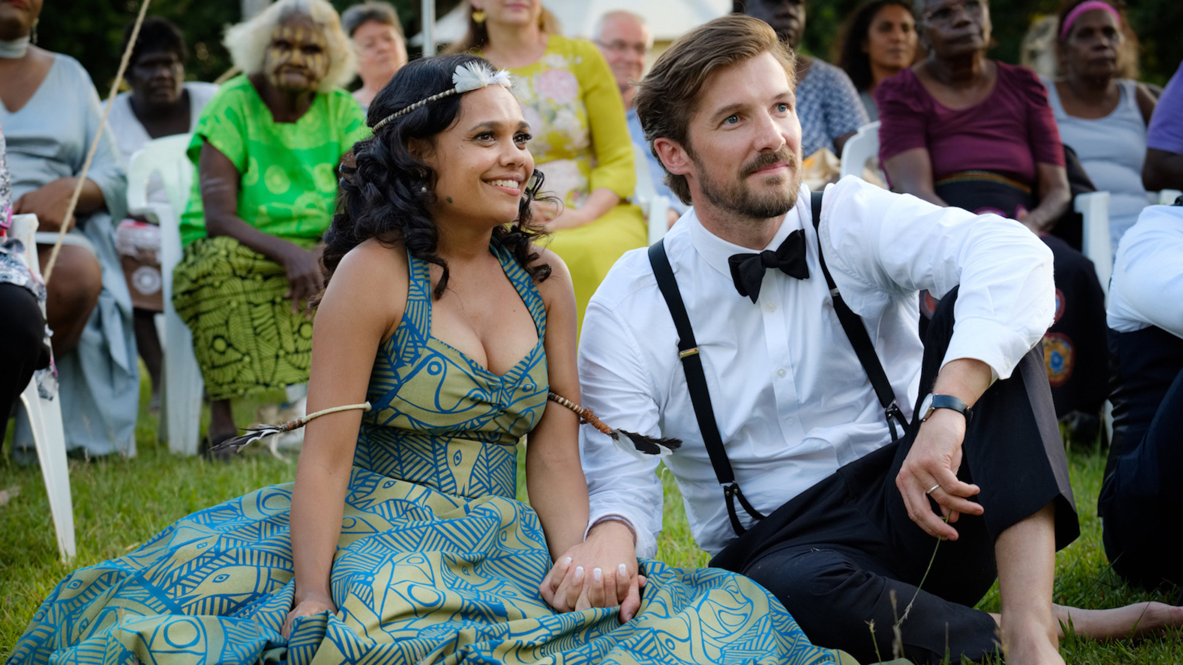 "Meine chaotische Hochzeit": Lauren (Miranda Tapsell) und Ned (Gwilym Lee) sitzen glücklich lächelnd, Hand in Hand, nebeneinander im Gras. Im Hintergrund sind weitere Gäste um das Paar versammelt.