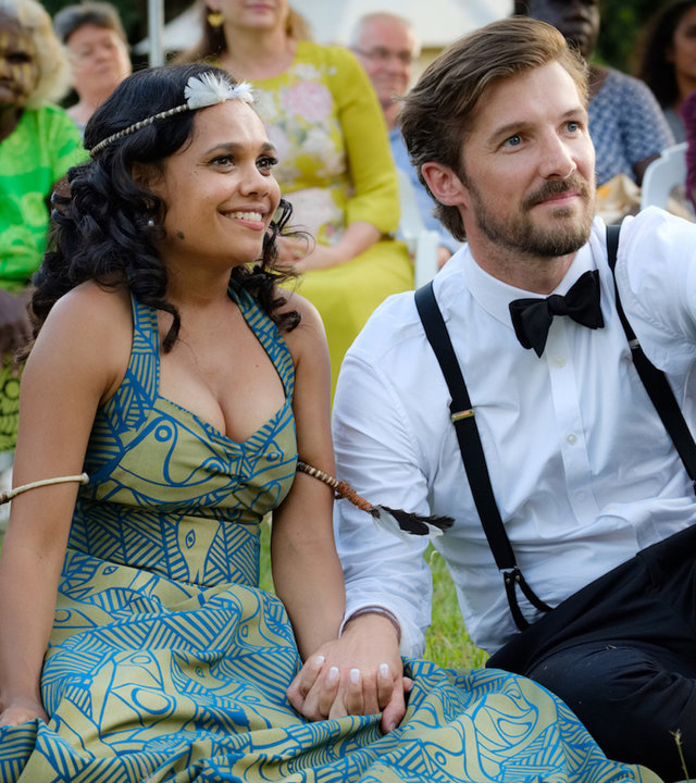 "Meine chaotische Hochzeit": Lauren (Miranda Tapsell) und Ned (Gwilym Lee) sitzen glücklich lächelnd, Hand in Hand, nebeneinander im Gras. Im Hintergrund sind weitere Gäste um das Paar versammelt.