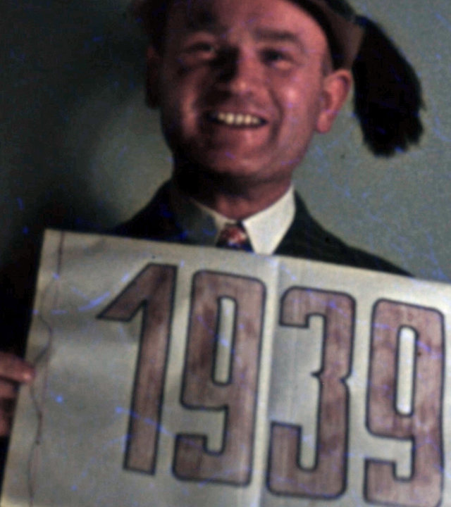 "Wir im Krieg – Privatfilme aus der NS-Zeit": Mann in Tracht hält Schild mit Jahreszahl 1939 hoch und lacht in die Kamera. "Wir im Krieg. Privatfilme aus der NS-Zeit".