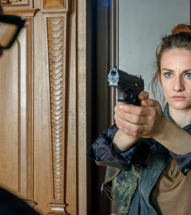 "SOKO Leipzig - Weihnachten im April": Natalie Kuczinski (Karoline Teska) zielt mit einer Waffe auf Richter Paffner (Thorsten Giese), der vor ihr steht. Sie steht mit dem Rücken an einer geschlossenen Holztür. Ihr Blick ist ernst.