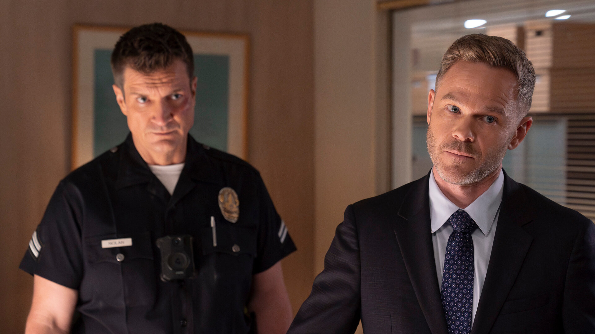 "The Rookie - Der Kuss": Officer John Nolan (Nathan Fillion) und Wesley Evers (Shawn Ashmore) stehen nebeneinander in einem Büroraum. Nolan trägt eine dunkle, kurzärmlige Dienstuniform und blickt ernst nach vorn. Wesley trägt einen schwarzen Anzug und schaut nachdenklich zur Seite.