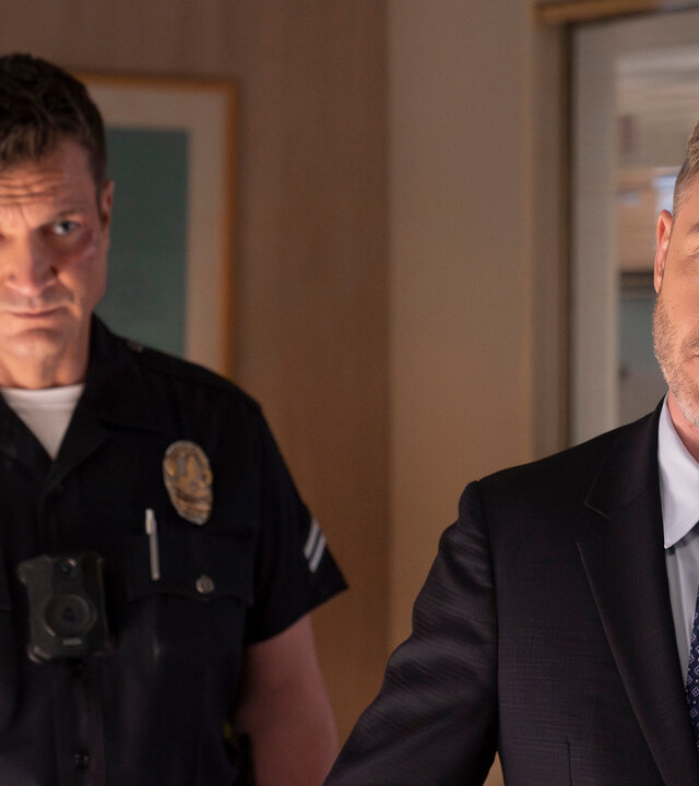"The Rookie - Der Kuss": Officer John Nolan (Nathan Fillion) und Wesley Evers (Shawn Ashmore) stehen nebeneinander in einem Büroraum. Nolan trägt eine dunkle, kurzärmlige Dienstuniform und blickt ernst nach vorn. Wesley trägt einen schwarzen Anzug und schaut nachdenklich zur Seite.