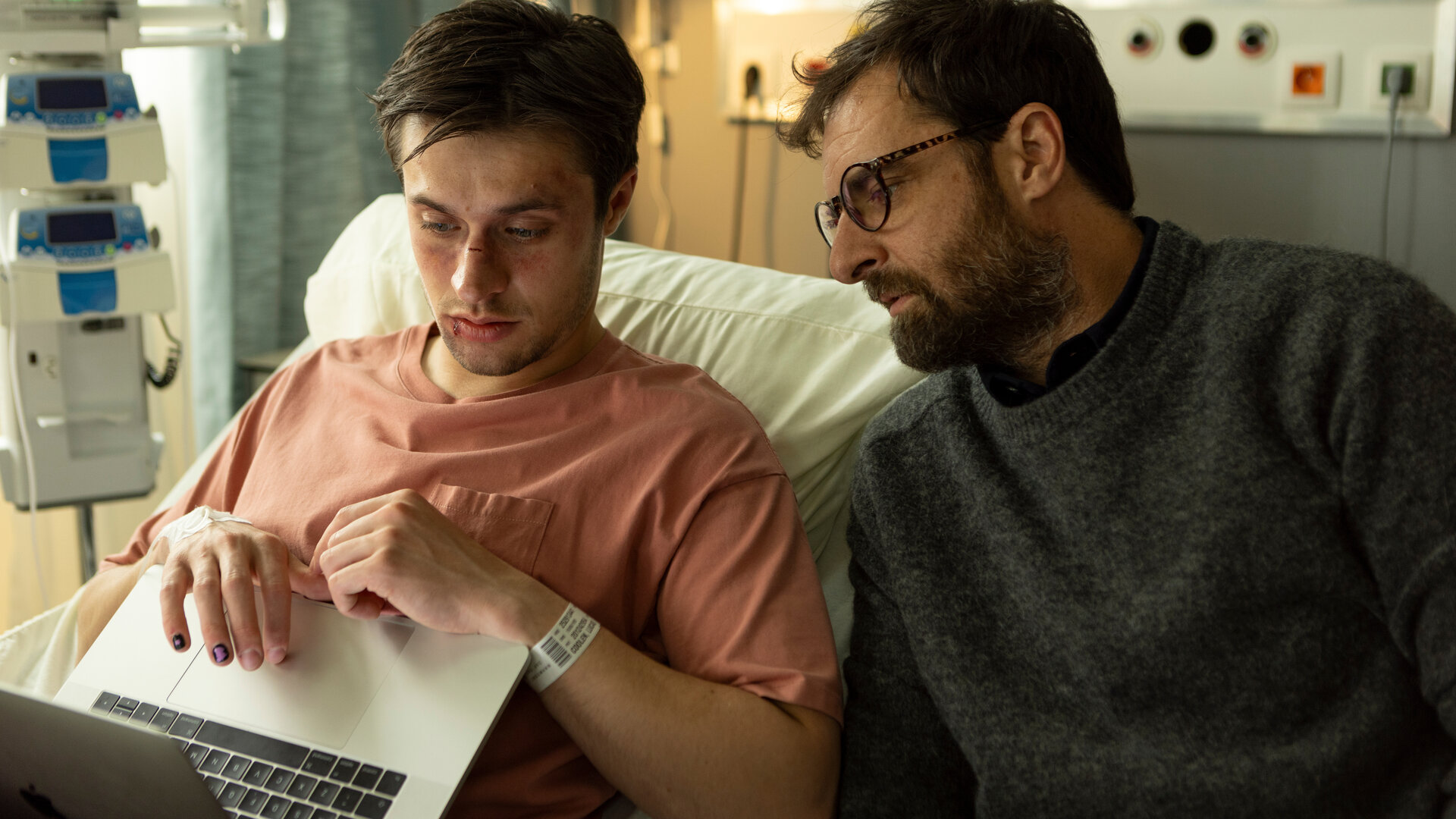 "The Kollective - Wir sind Millionen": Lucas (Gijs Blom) sitzt aufrecht im Krankenbett und blickt auf seinen aufgeklappten Laptop. Neben ihm sitzt Etienne (Grégory Montel) und schaut ebenfalls in die Richtung des Computers.