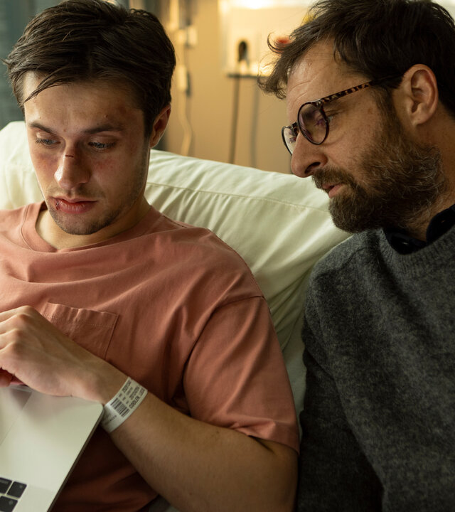 "The Kollective - Wir sind Millionen": Lucas (Gijs Blom) sitzt aufrecht im Krankenbett und blickt auf seinen aufgeklappten Laptop. Neben ihm sitzt Etienne (Grégory Montel) und schaut ebenfalls in die Richtung des Computers.