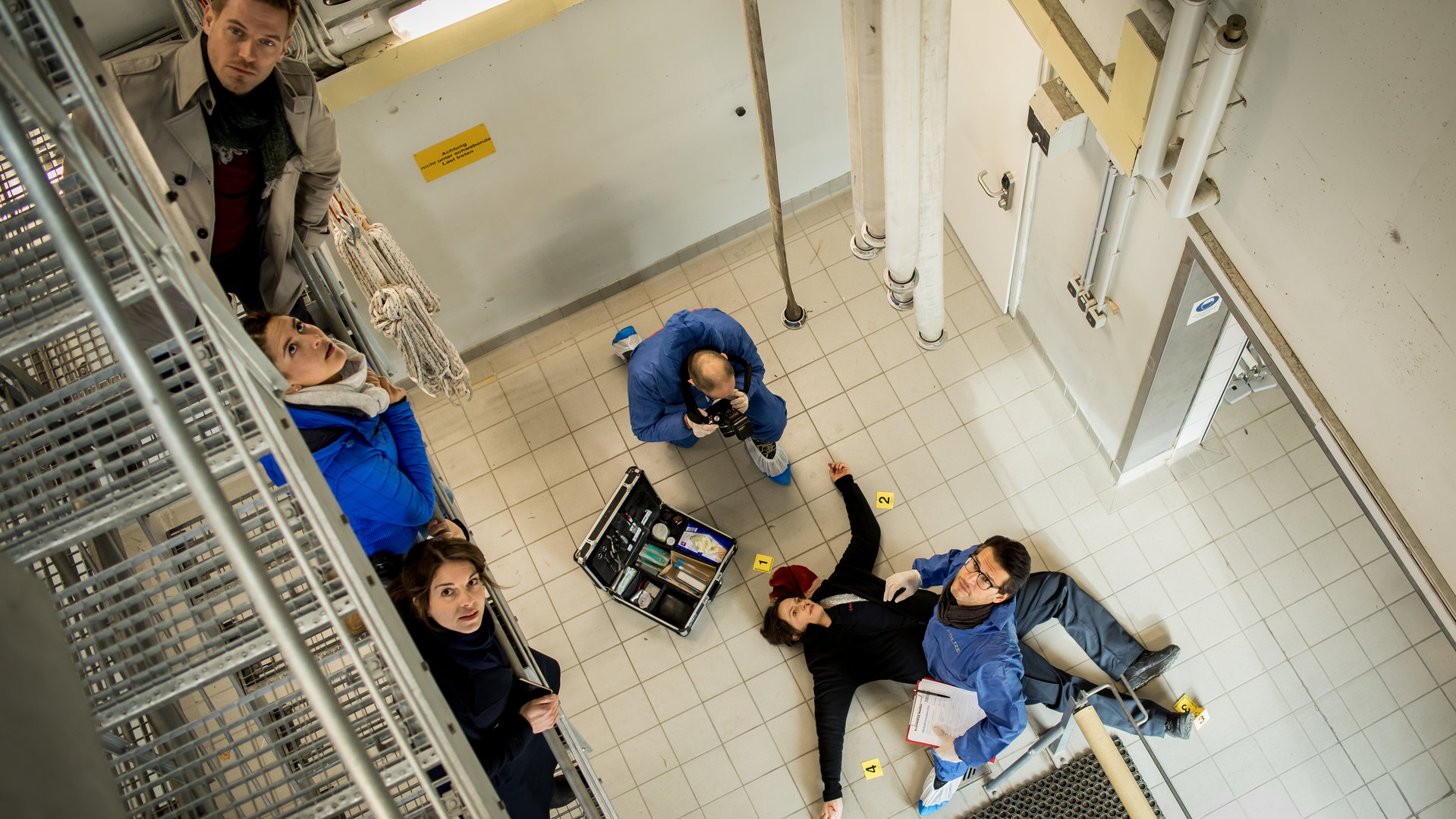 "SOKO München - Feuerland": Blick von oben auf die Szene: Auf der Wache der Feuerwehr liegt eine Frau tot auf dem Boden, unter ihrem Kopf ist Blut zu sehen. Neben ihr steht Dr. Weissenböck (Florian Odendahl) mit Unterlagen in der Hand, er schaut nach oben zur die Treppe. Am Kopf der Leiche steht ein Koffer mit Untersuchungsutensilien, daneben sitzt ein Mann von der Spusi in der Hocke und macht Beweisfotos von der Leiche. Auf der Treppe stehen Dominik Morgenstern (Joscha Kiefer), Toni Bischoff (Amanda da Gloria) und Katharina Hahn (Bianca Hein), sie schauen hoch zur nächsten Treppenetage.