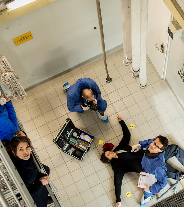 "SOKO München - Feuerland": Blick von oben auf die Szene: Auf der Wache der Feuerwehr liegt eine Frau tot auf dem Boden, unter ihrem Kopf ist Blut zu sehen. Neben ihr steht Dr. Weissenböck (Florian Odendahl) mit Unterlagen in der Hand, er schaut nach oben zur die Treppe. Am Kopf der Leiche steht ein Koffer mit Untersuchungsutensilien, daneben sitzt ein Mann von der Spusi in der Hocke und macht Beweisfotos von der Leiche. Auf der Treppe stehen Dominik Morgenstern (Joscha Kiefer), Toni Bischoff (Amanda da Gloria) und Katharina Hahn (Bianca Hein), sie schauen hoch zur nächsten Treppenetage.
