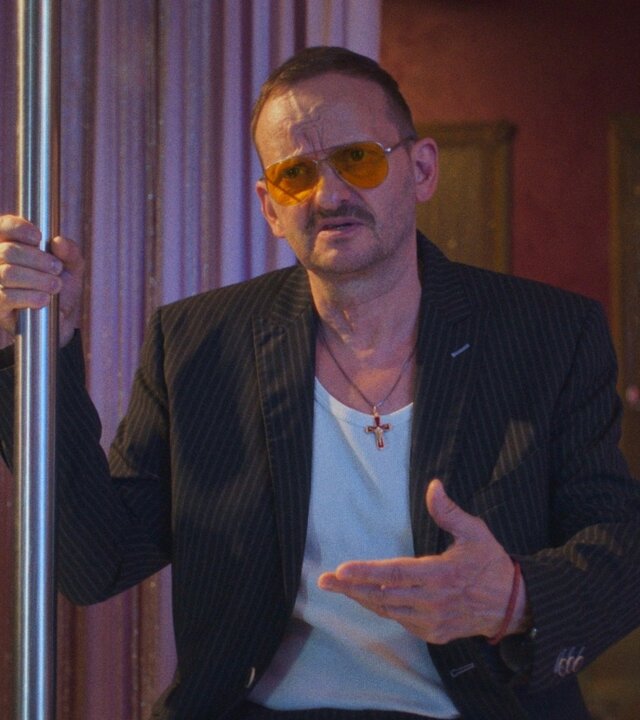 "Doppelhaushälfte – Erben": Andi Knuppe (Milan Peschel) steht in einem Clubraum an einer Stange, trägt Sonnenbrille und Anzug. Er gestikuliert selbstbewusst, buntes Licht beleuchtet die Szene.