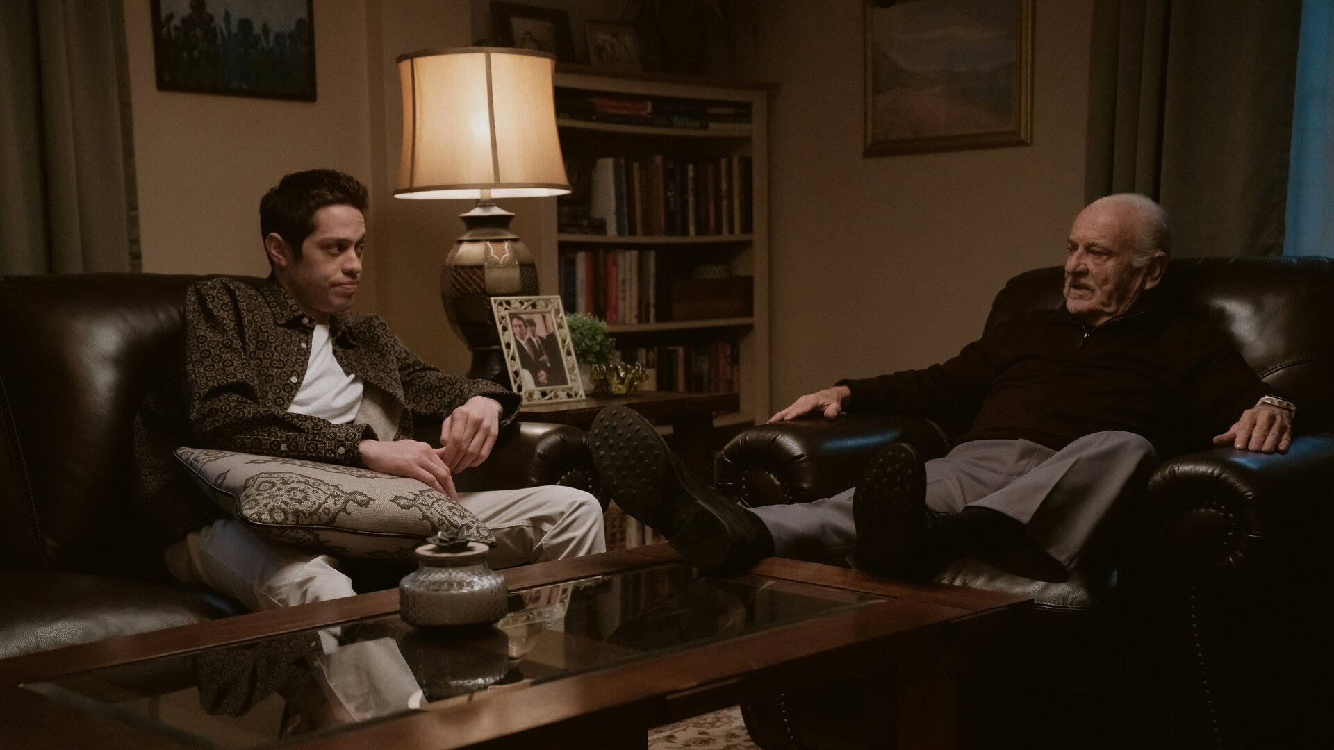 "Bupkis - Die Wandlung": Pete (Pete Davidson) sitzt auf einem Ledersofa. Auf seinem Schoß liegt ein Kissen. Er schaut Opa Joe (Joe Pesci) an, der im Ledersessel sitzt und mit Pete redet. Beide wirken sehr ernst. Der Raum ist dunkel, es leuchtet nur eine Tischlampe und die Stimmung wirkt bedrückend.