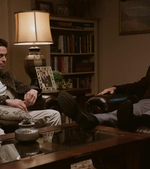 "Bupkis - Die Wandlung": Pete (Pete Davidson) sitzt auf einem Ledersofa. Auf seinem Schoß liegt ein Kissen. Er schaut Opa Joe (Joe Pesci) an, der im Ledersessel sitzt und mit Pete redet. Beide wirken sehr ernst. Der Raum ist dunkel, es leuchtet nur eine Tischlampe und die Stimmung wirkt bedrückend.