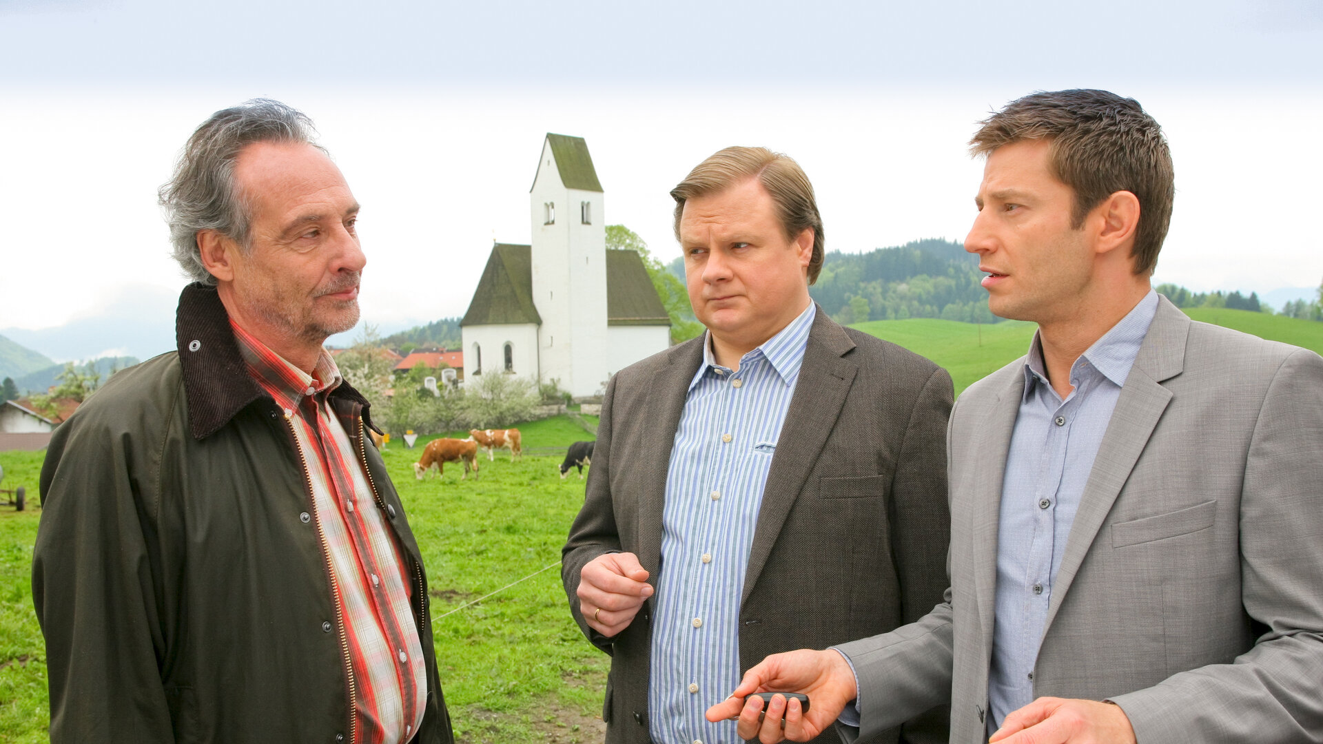 "Die Rosenheim-Cops - Sprung in den Tod": Auf einer Kuhweide an einer Kirche befragen die Ermittler Hansen (Igor Jeftic) und Hartl (Michael A. Grimm) Tierarzt Sendlinger (Daniel Friedrich).