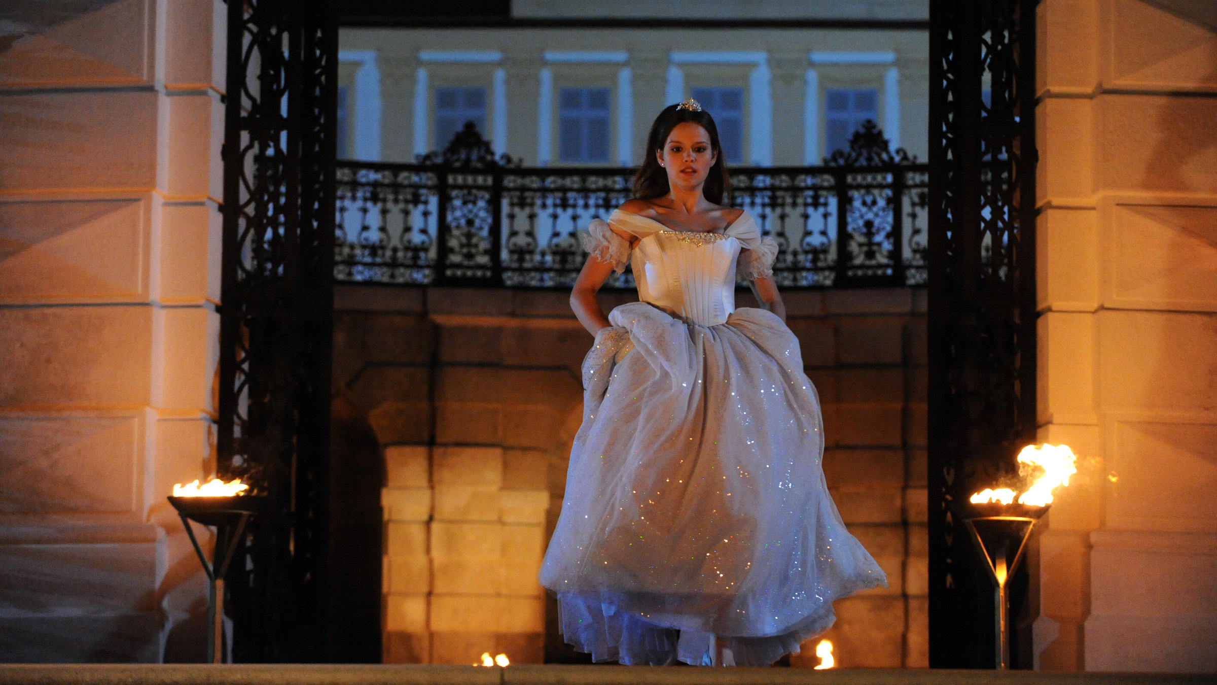 "Aschenputtel": Im Hintergrund ist das nächtliche Schloss zu sehen. Aschenputtel (Emilia Schüle) hat ihr Kleid gerafft und eilt durch das eiserne Eingangstor. Zu den Seiten des Tores steht jeweils eine Fackel.