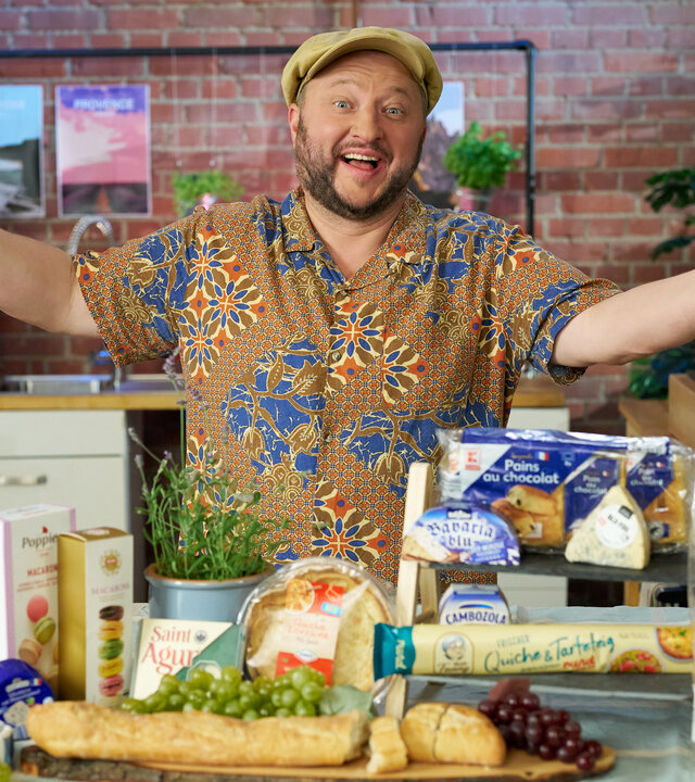 "besseresser: Freche Frankreich-Fakes": Sebastian Lege steht hinter französischen Produkten, die auf einem Tisch liegen, und lächelt in die Kamera.