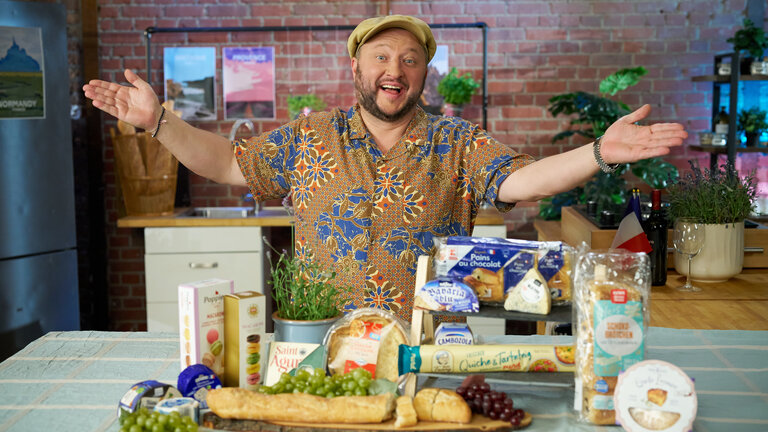 "besseresser: Freche Frankreich-Fakes": Sebastian Lege steht hinter französischen Produkten, die auf einem Tisch liegen, und lächelt in die Kamera.