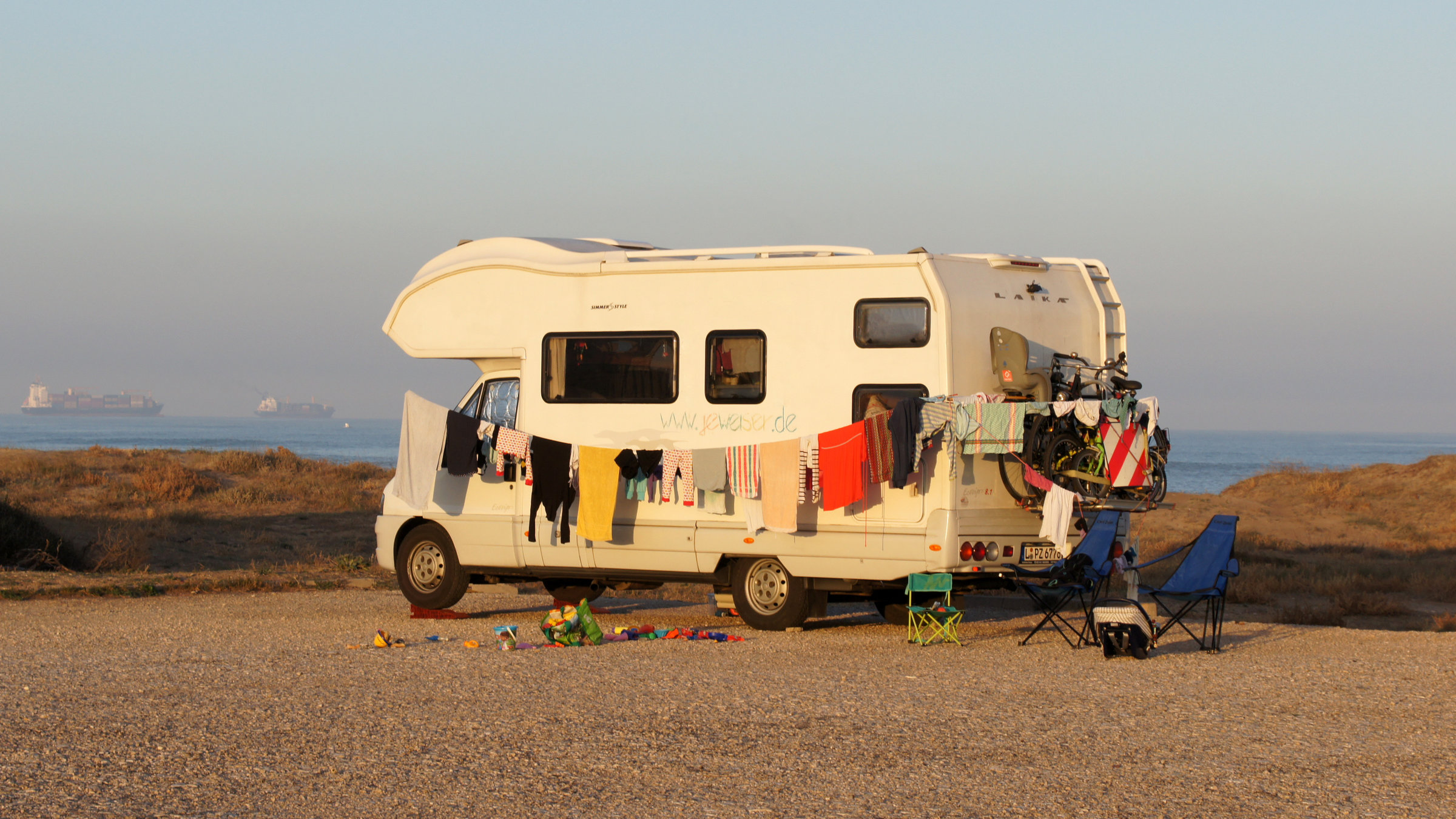"37°: Mein Haus zieht mit - Abenteuer Hausboot, Camper, Tiny House": Der Camper steht allein auf einem Parkplatz, im Hintergrund sieht man das Meer. Entlang des Campers ist eine Wäscheleine gespannt, an der bunte Wäschestücke hängen.