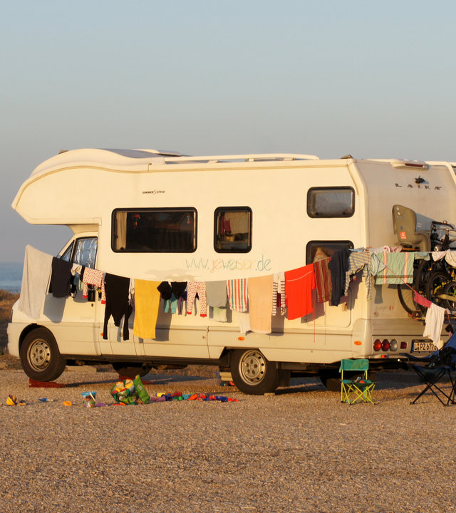 "37°: Mein Haus zieht mit - Abenteuer Hausboot, Camper, Tiny House": Der Camper steht allein auf einem Parkplatz, im Hintergrund sieht man das Meer. Entlang des Campers ist eine Wäscheleine gespannt, an der bunte Wäschestücke hängen.