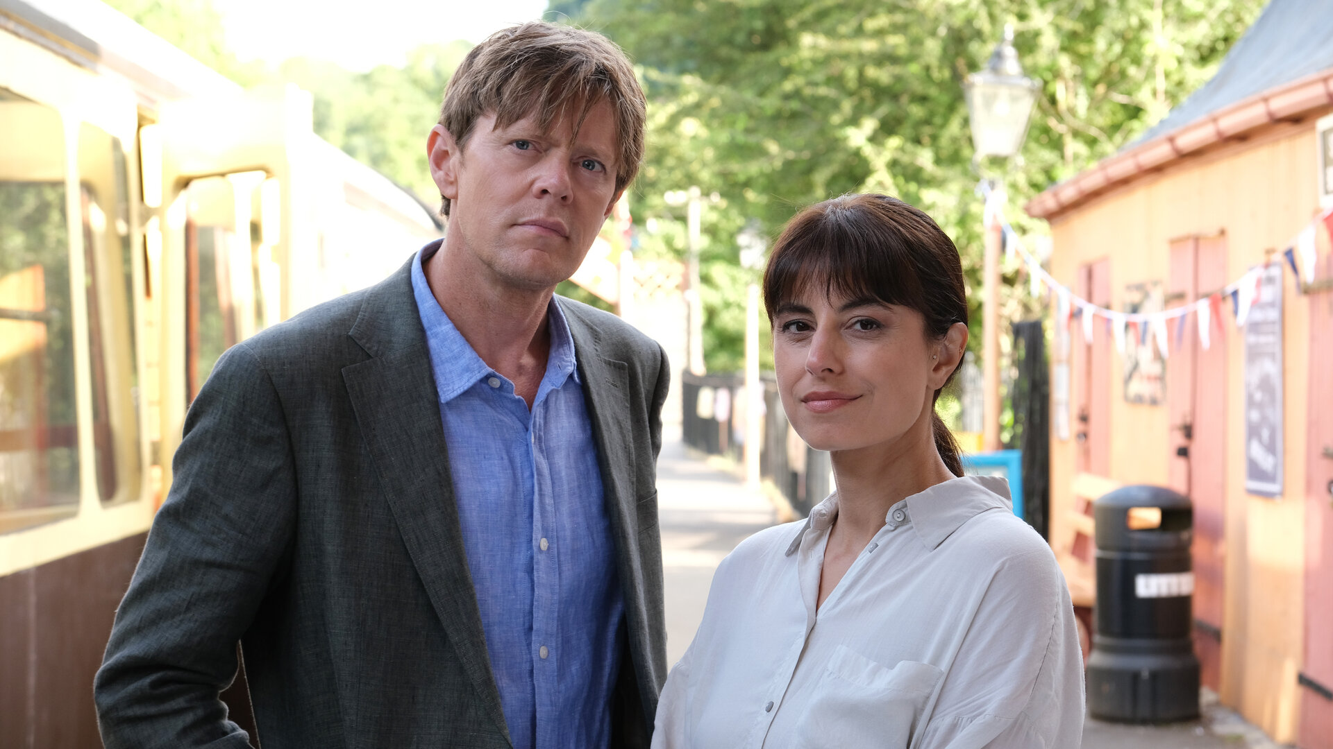 "Beyond Paradise - Mord im Theaterexpress": DI Humphrey Goodman (Kris Marshall) und DS Esther Williams (Zahra Ahmadi) stehen nebeneinander auf einem Bahnsteig und schauen interessiert nach vorne. Im Hintergrund links hält ein Passagierwaggon, rechts steht ein niedriges Bahnhofsgebäude.