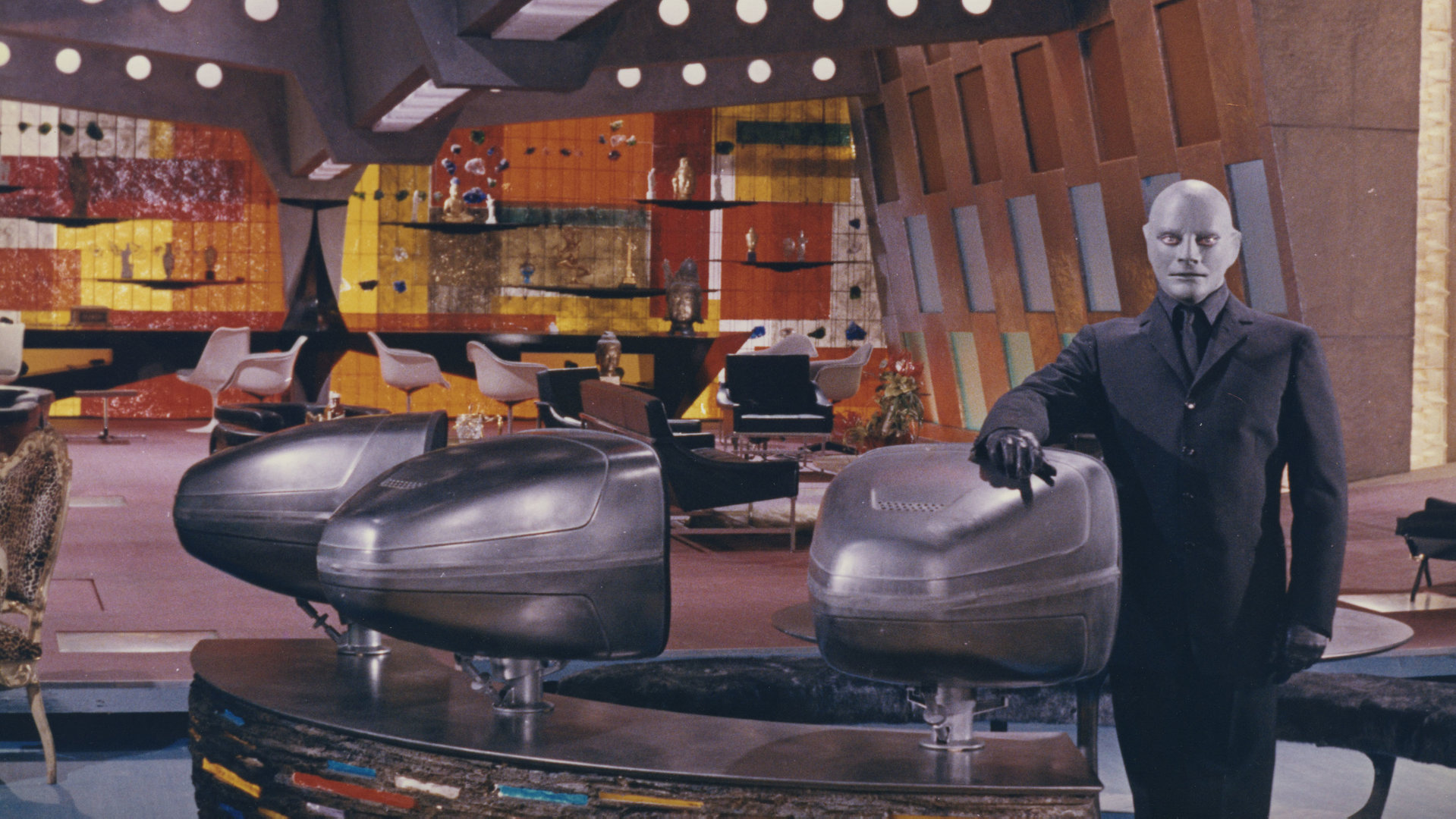 "Fantomas gegen Interpol": Fantomas (Jean Marais) lehnt an einen Fernseher in einem hell erleuchteten Raum und schaut mit ernster Miene nach vorne. Futuristische Gerät stehen in dem Raum.