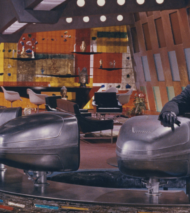 "Fantomas gegen Interpol": Fantomas (Jean Marais) lehnt an einen Fernseher in einem hell erleuchteten Raum und schaut mit ernster Miene nach vorne. Futuristische Gerät stehen in dem Raum.