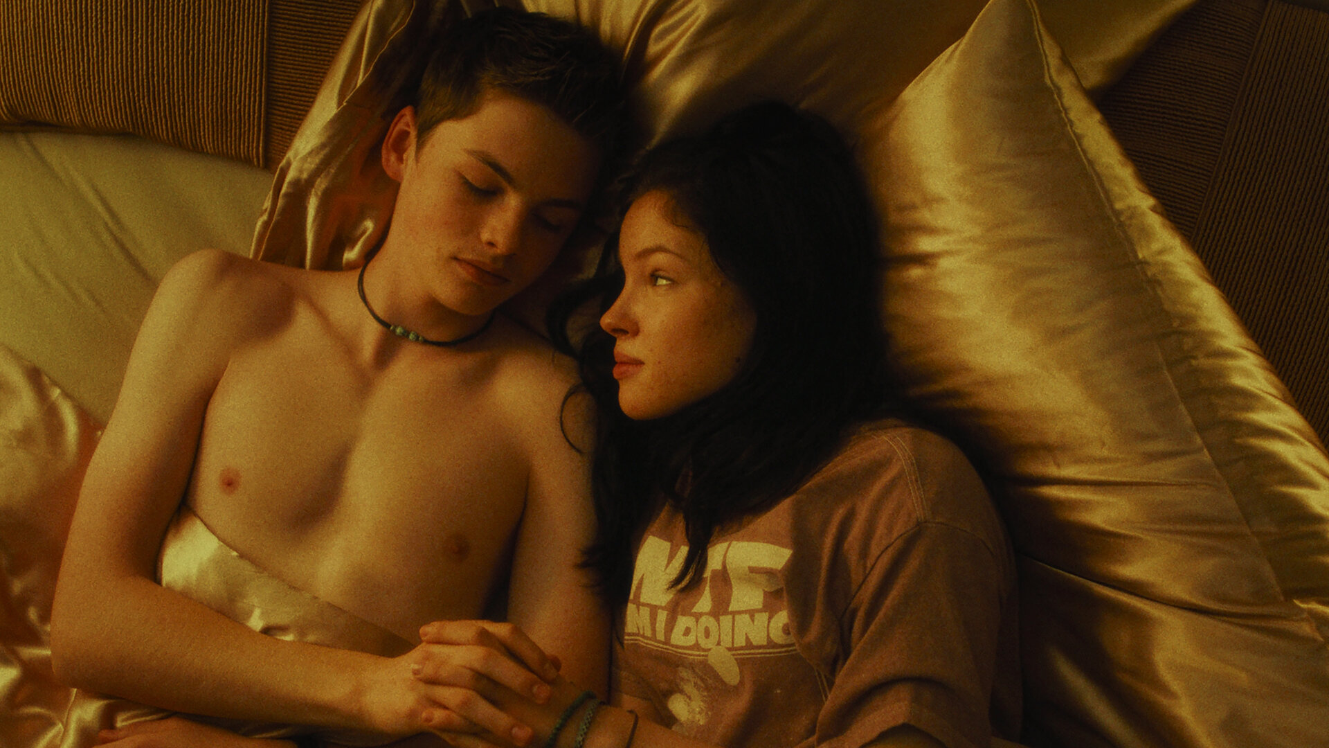 "Chabos - Du bist Deutschland": Blick von schräg oben: Peppi (Nico Marischka), mit nacktem Oberkörper, und Mascha (Arina Prass) liegen zugedeckt mit goldfarbener Bettwäsche nebeneinander im Bett. Sie halten Händchen und schauen einander an.