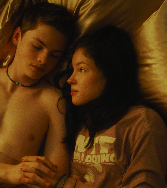 "Chabos - Du bist Deutschland": Blick von schräg oben: Peppi (Nico Marischka), mit nacktem Oberkörper, und Mascha (Arina Prass) liegen zugedeckt mit goldfarbener Bettwäsche nebeneinander im Bett. Sie halten Händchen und schauen einander an.