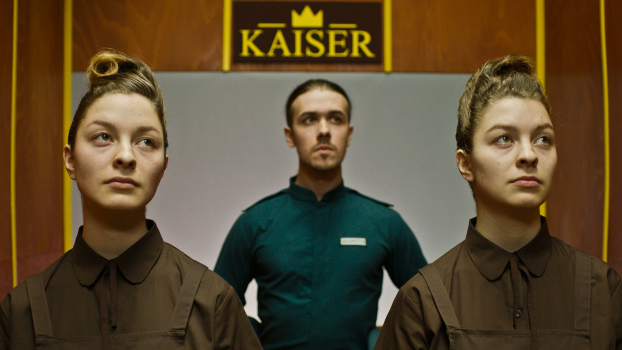 "KlassiXS: Macht und Hierarchien - Kaiser": Zwei Frauen und ein Mann im Hintergrund, die Frauen sind Zwillinge und haben die gleiche Frisur und das gleiche braune Oberteil an. Der Mann im Hintergrund hat die Haare zusammengebunden und trägt ein grünes Hemd. An der Wand hinter ihnen ist ein Logo zu sehen, auf dem „Kaiser“ steht.