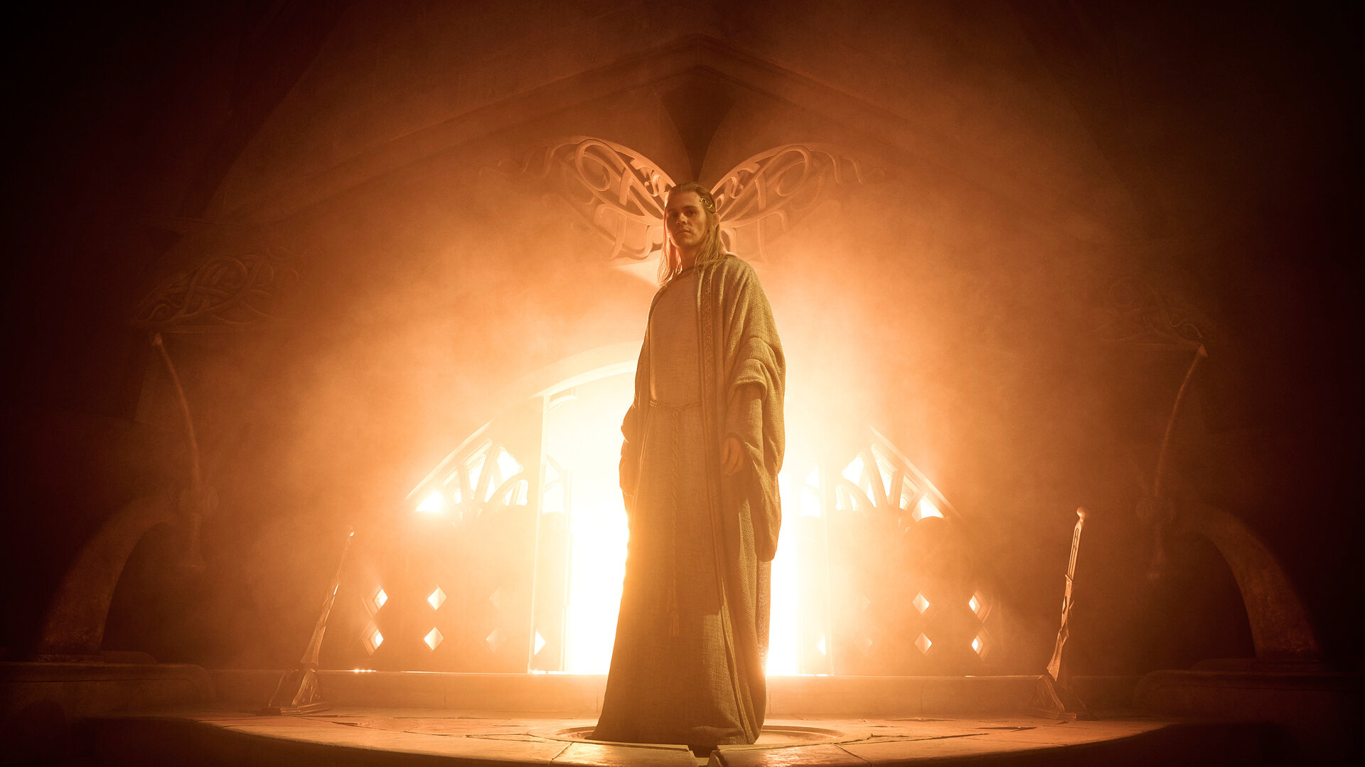 "Der Herr der Ringe: Die Ringe der Macht - Wo die Sterne fremd sind": Sauron (Charlie Vickers) steht in einem dunklen Raum vor einer großen Feuerstelle. Er trägt lange Haare und ein helles Gewand.