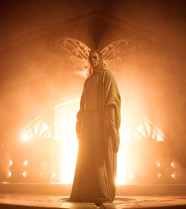 "Der Herr der Ringe: Die Ringe der Macht - Wo die Sterne fremd sind": Sauron (Charlie Vickers) steht in einem dunklen Raum vor einer großen Feuerstelle. Er trägt lange Haare und ein helles Gewand.