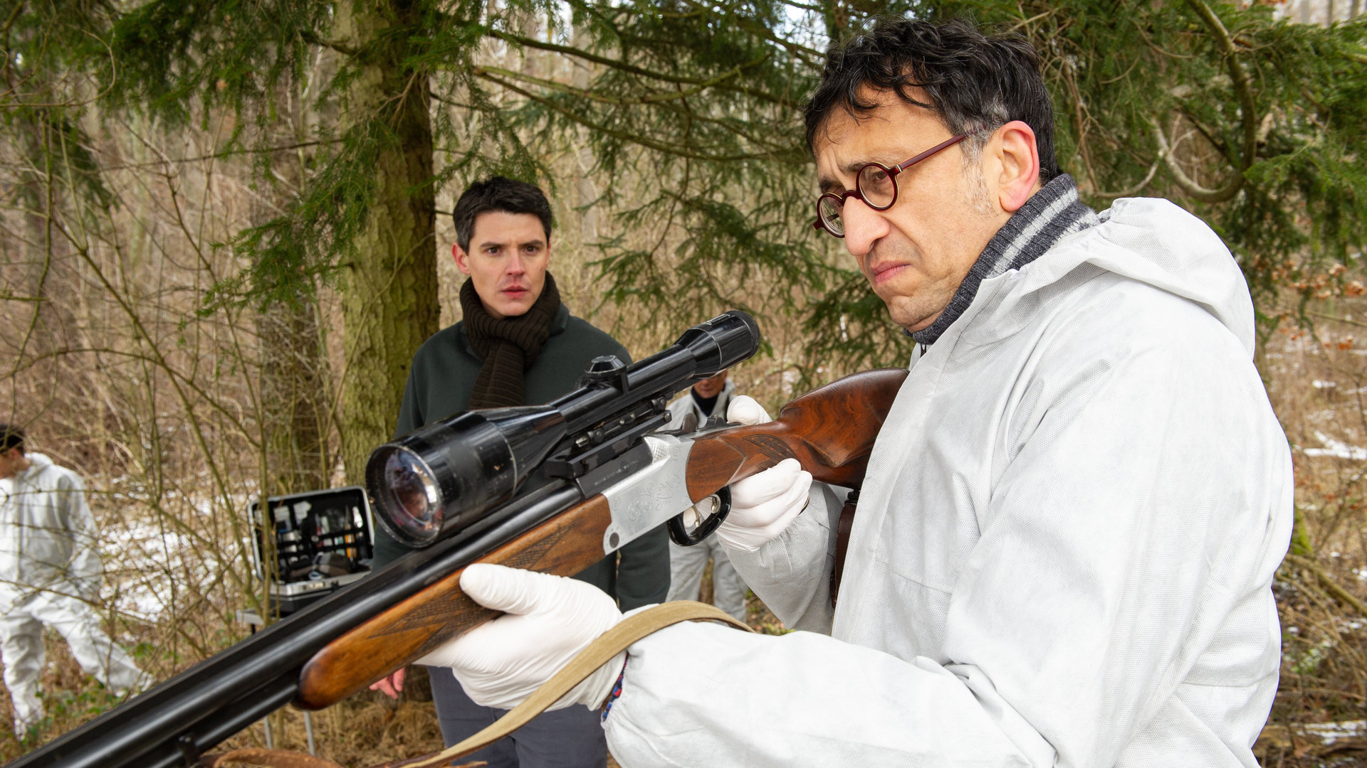 "SOKO Stuttgart - Böser Wolf": Im winterlichen Wald untersucht Jan Arnaud (Mike Zaka Sommerfeldt) im weißen Spurensicherungsanzug ein Jagdgewehr. Er wird von Rico Sander (Benjamin Strecker) dabei beobachtet, wie er das Gewehr in Schussposition hält. Im Hintergrund sind zwei Mitarbeiter der Spurensicherung (Komparsen) am Arbeiten.