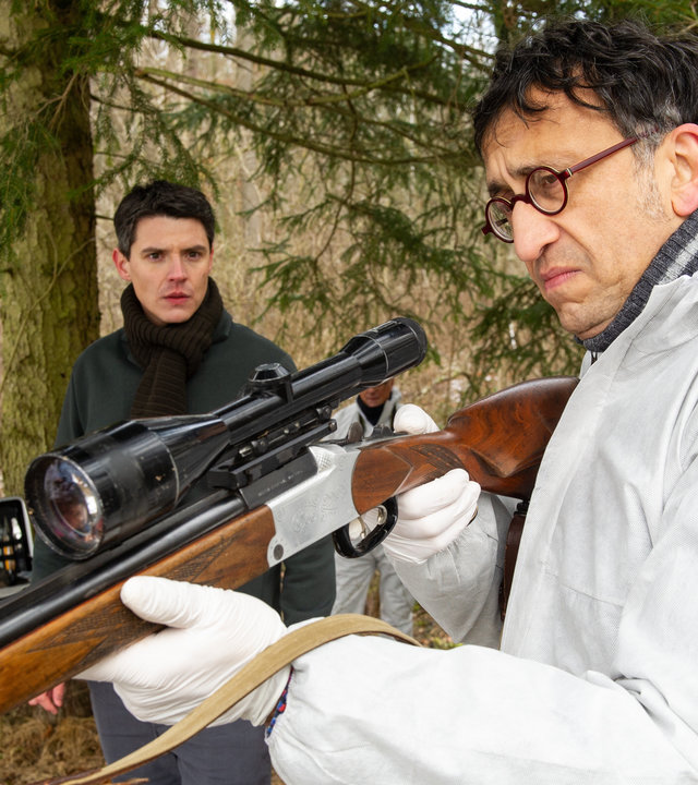 "SOKO Stuttgart - Böser Wolf": Im winterlichen Wald untersucht Jan Arnaud (Mike Zaka Sommerfeldt) im weißen Spurensicherungsanzug ein Jagdgewehr. Er wird von Rico Sander (Benjamin Strecker) dabei beobachtet, wie er das Gewehr in Schussposition hält. Im Hintergrund sind zwei Mitarbeiter der Spurensicherung (Komparsen) am Arbeiten.