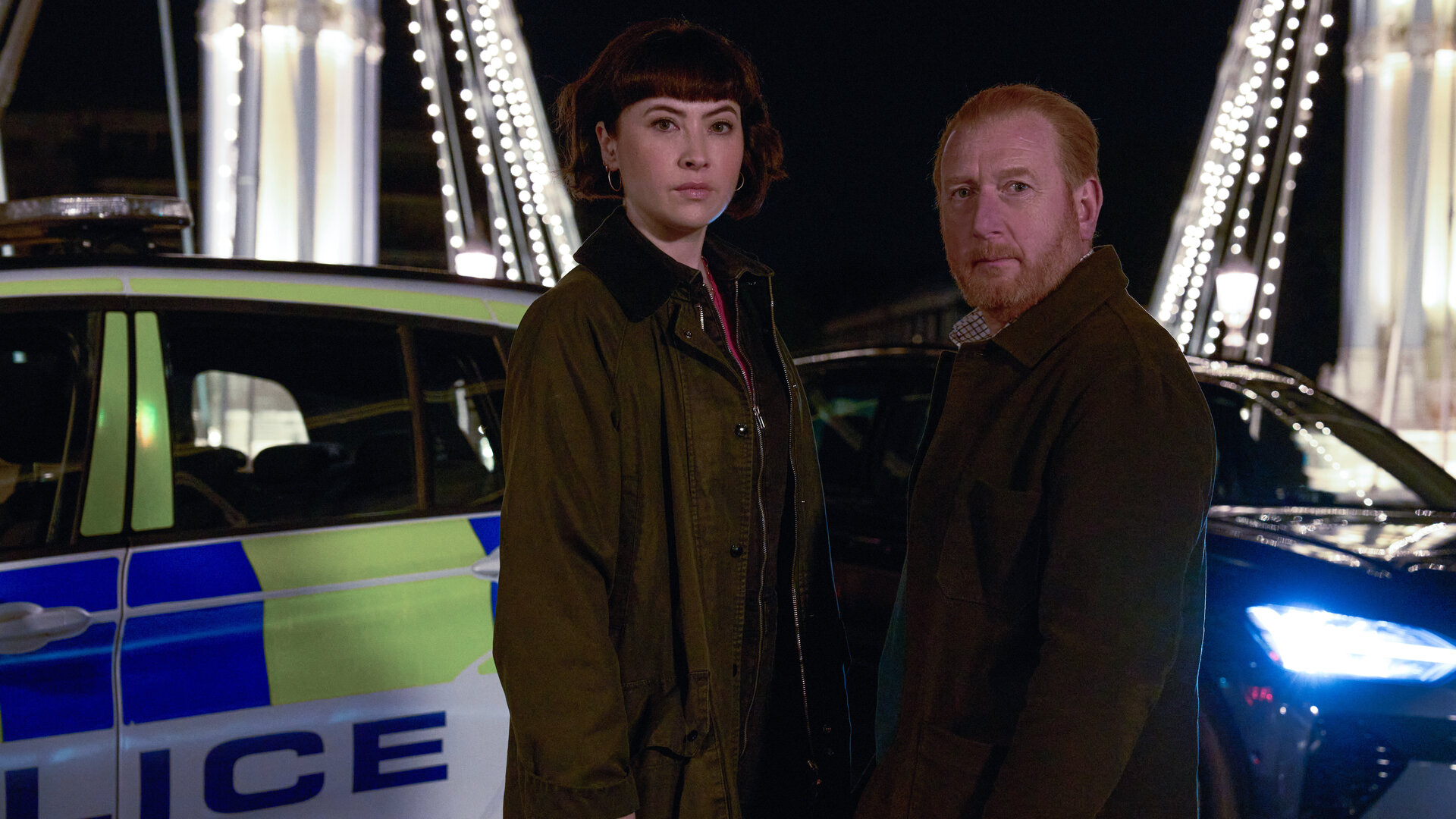 "The Chelsea Detective - Alle liebten Chloe": DI Max Arnold (Adrian Scarborough) und DS Layla Walsh (Vanessa Emme) stehen nachts nebeneinander auf einer Brücke in London, deren Pfeiler durch zahlreichen Glühlampen beleuchtet werden, und schauen in die Kamera. Hinter ihnen versperren ein Streifenwagen und ein ziviles Auto die Straße.