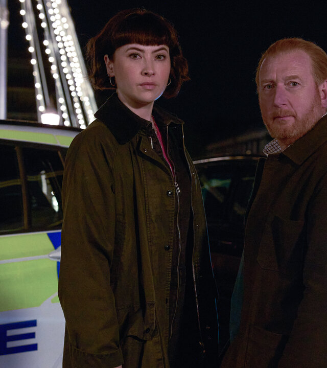 "The Chelsea Detective - Alle liebten Chloe": DI Max Arnold (Adrian Scarborough) und DS Layla Walsh (Vanessa Emme) stehen nachts nebeneinander auf einer Brücke in London, deren Pfeiler durch zahlreichen Glühlampen beleuchtet werden, und schauen in die Kamera. Hinter ihnen versperren ein Streifenwagen und ein ziviles Auto die Straße.