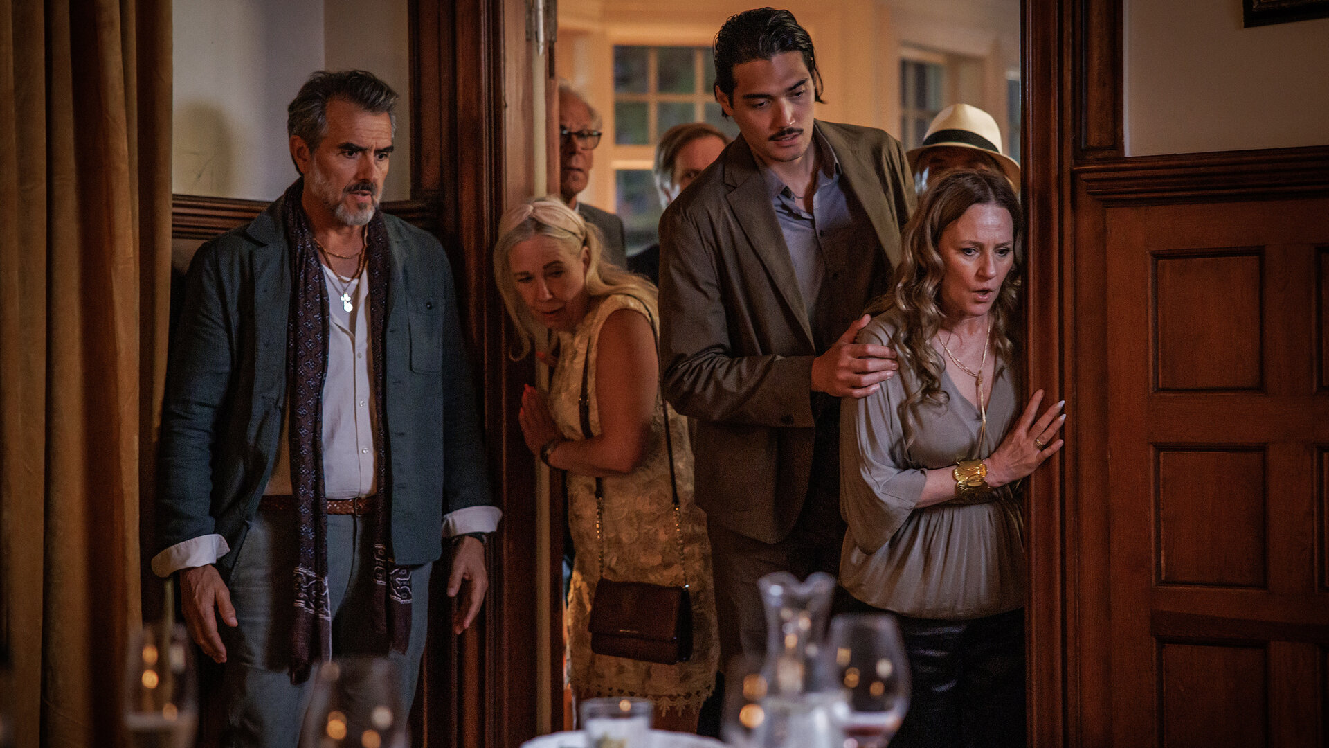 "Mord im Mittsommer - Linnea": Isidor (Rafael Edholm), Edvin (Adam Dalström) und Catharina (Annika Hallin) stehen nebeneinander in einem Raum mit holzvertäfelten Wänden. Sie beobachten etwas. Hinter ihnen ist ein Durchgang zu einem weiteren Zimmer, in dem sich mehrere Menschen (Komparsen) aufhalten.