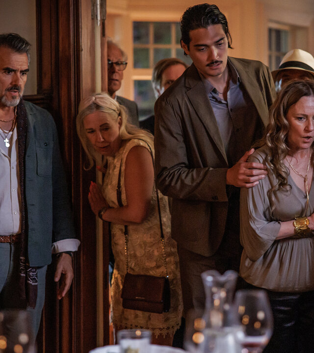 "Mord im Mittsommer - Linnea": Isidor (Rafael Edholm), Edvin (Adam Dalström) und Catharina (Annika Hallin) stehen nebeneinander in einem Raum mit holzvertäfelten Wänden. Sie beobachten etwas. Hinter ihnen ist ein Durchgang zu einem weiteren Zimmer, in dem sich mehrere Menschen (Komparsen) aufhalten.