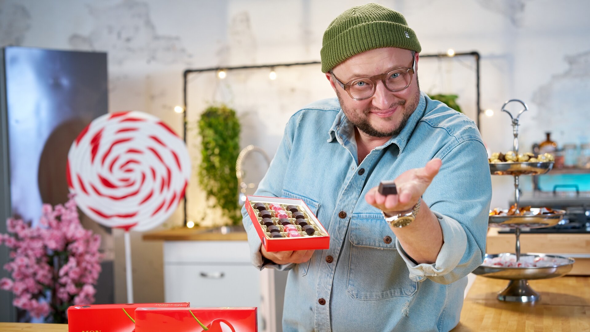 "besseresser: Die Wahrheit über Ferrero - Food Stories mit Sebastian Lege": Sebastian Lege hält eine geöffnete Packung Mon Chérie in der rechten Hand und präsentiert eine Schokopraline mit der linken Hand.