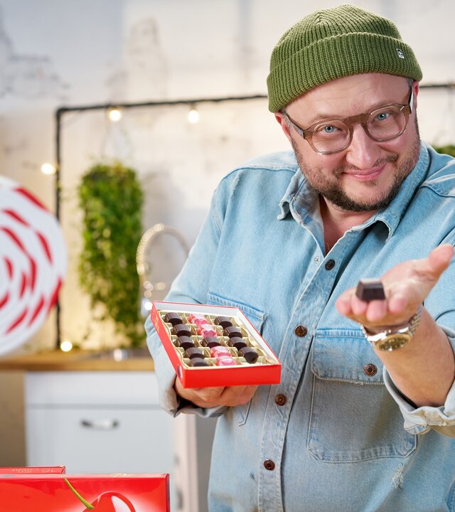 "besseresser: Die Wahrheit über Ferrero - Food Stories mit Sebastian Lege": Sebastian Lege hält eine geöffnete Packung Mon Chérie in der rechten Hand und präsentiert eine Schokopraline mit der linken Hand.
