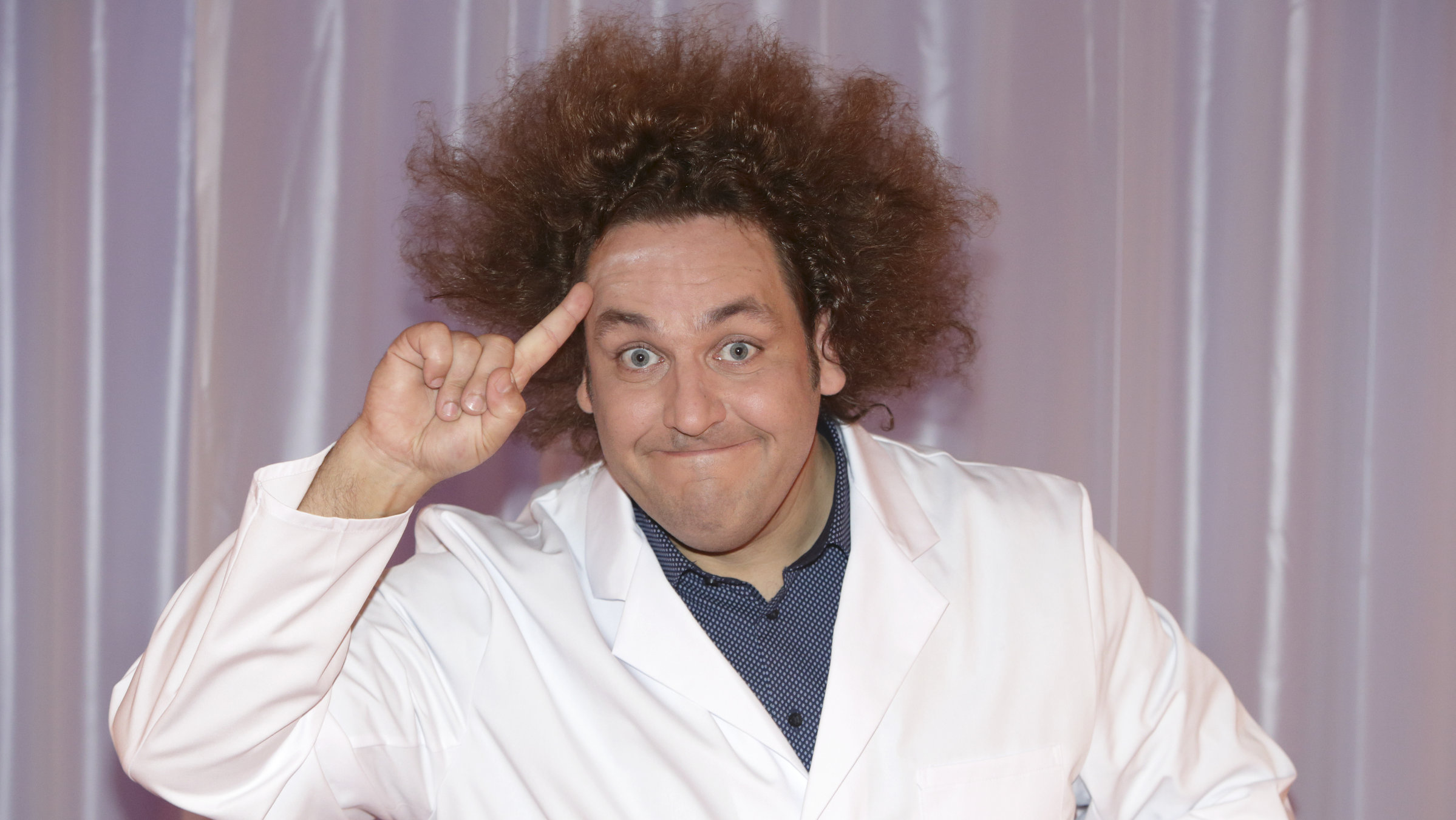 "Konrad Stöckel: Wie man mit AC/DC das Licht ausmacht": Konrad Stöckel im weißen Kittel mit erhobenen Zeigefinger an der Stirn. Er schaut grinsend in die Kamera.