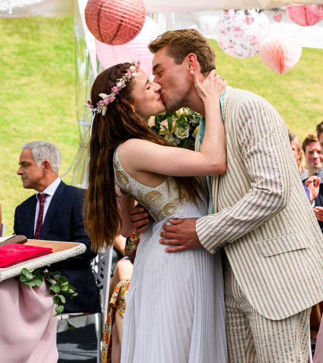 "Bettys Diagnose - Hochzeitsfieber": Die Standesbeamtin (Hella-Birgit Mascus) steht in dunkelgrünem Kleid am Stehpult und sieht Sofia (Kiara Brunken) und Lukas (Niklas Löffler) an, die sich küssen. Im Hintergrund sitzt die Hochzeitsgesellschaft auf Stühlen und applaudiert lächelnd.