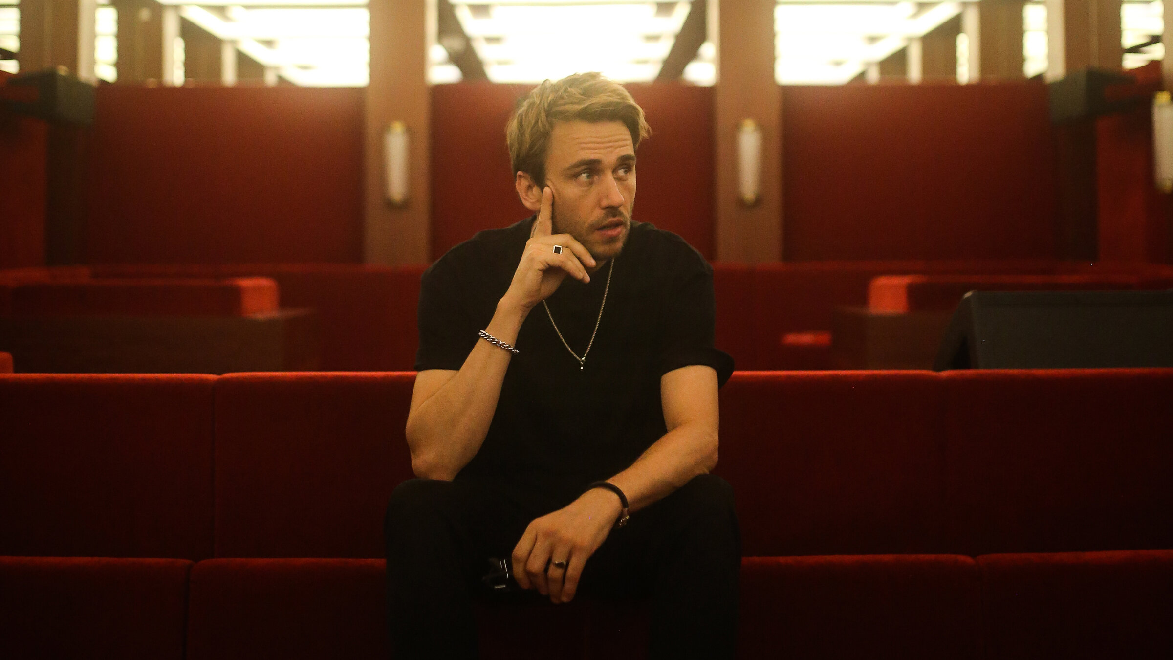 "Clueso: Sommer Tour 2024 - Live von der Berliner Waldbühne" Der Pop-Künstler Clueso sitzt in einem Raum mit hell erleuchteter Decke und mit rotem Samt bezogenen Sitzbänken. Er selbst trägt eine schwarze Hose und T-Shirt und weiße Sneaker. Um die Handgelenke trägt er jeweils ein Armband, um den Hals eine Kette. Er hält eine Hand an sein Kinn und schaut zur Seite.