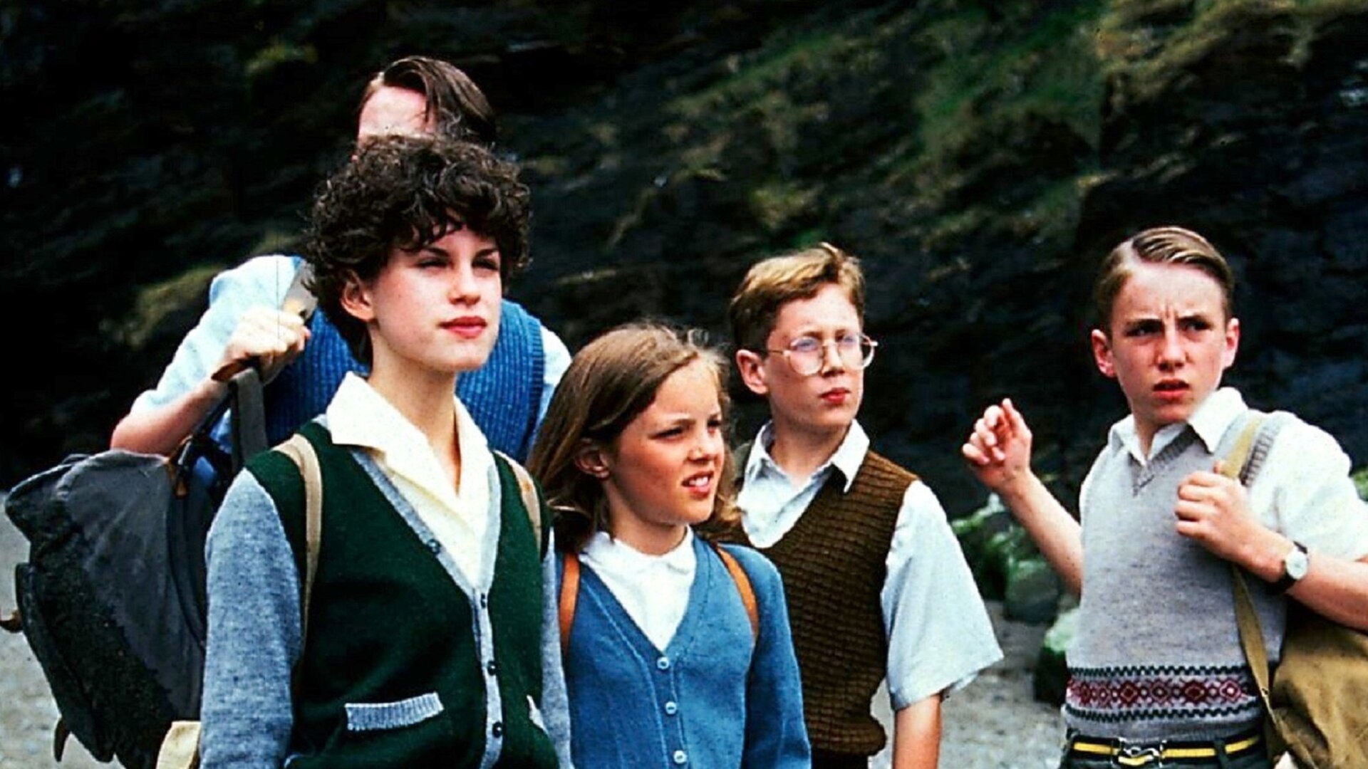 "Fünf Freunde verfolgen die Strandräuber": Julian (Marco Williamson), George (Jemima Rooper), Anne (Laura Petela), ein Junge und Dick (Paul Child) stehen nebeneinander vor einer steilen Felswand. Die Kinder schauen alle in eine Richtung.
