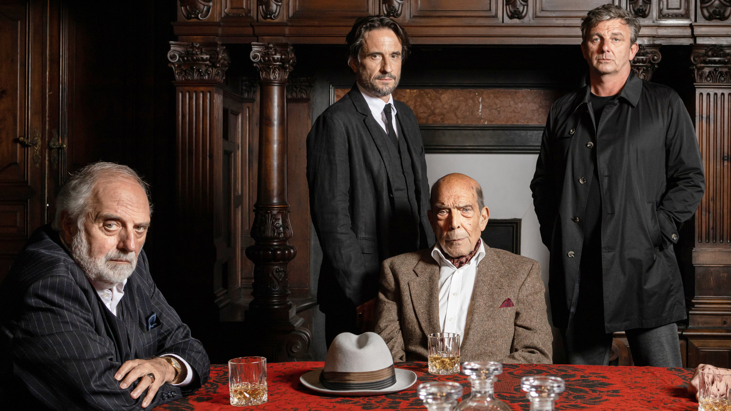 "Der Feind meines Feindes": Pietro Connagi (Alessandro Bressanello) und Toto Conte (Orso Maria Guerrini) sitzen an einem rot-schwarzen Tisch und blicken ernst in die Kamera. Auf dem Tisch stehen zwei halbvolle Whiskeygläser und zwischen diesen liegt ein grauer Hut. Hinter Toto Conte stehen Gabriel Morales (Oliver Mommsen) und Klaus Burg (Hans Sigl) nebeneinander und sehen ebenfalls in die Kamera. Klaus Burger trägt einen schwarzen Anorak, die anderen Männer auf dem Bild sind mit Anzügen bekleidet.