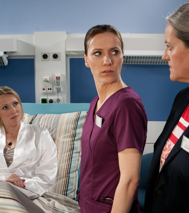 "Bettys Diagnose - Allein auf weitem Flur": Die Aufsichtsratsvorsitzende des Krankenhauses, Rebecca Ackermann (Dana Golombek) liegt im Krankenbett. Betty (Bettina Lamprecht) und Pflegedienstleiterin Mechthild Puhl (Sybille J. Schedwill) stehen neben dem Bett und werfen sich strenge Blicke zu.