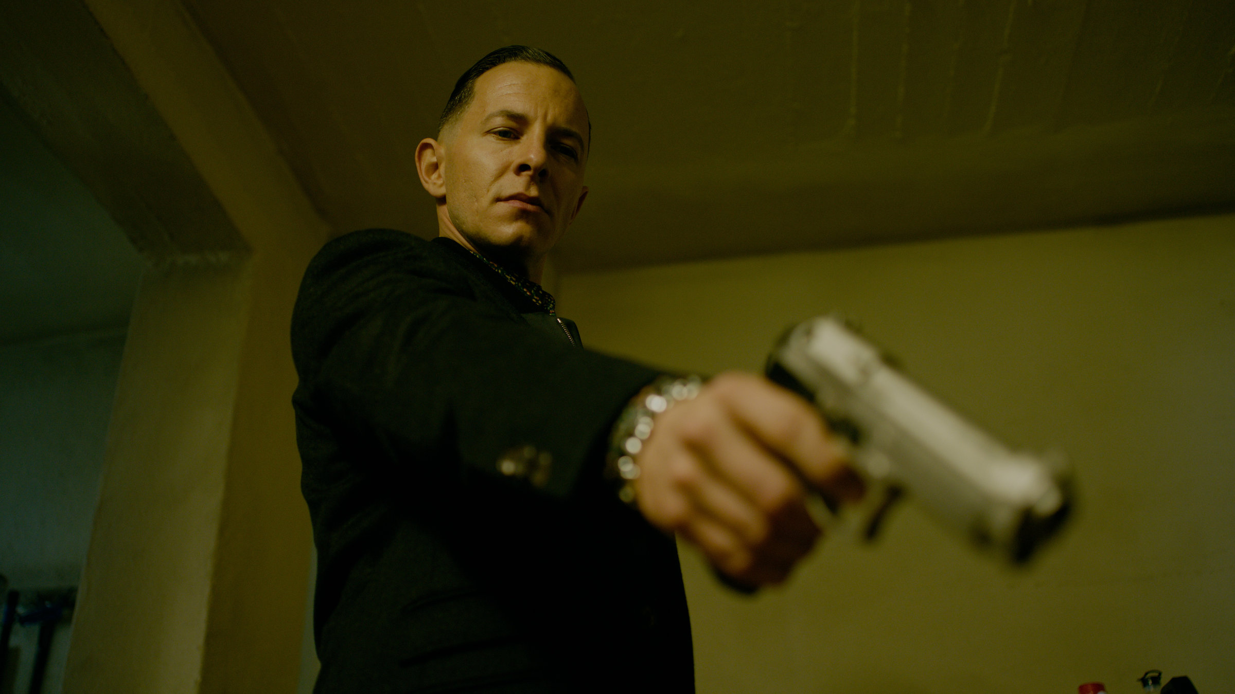 "Parfum - Fesselung": Unteransicht auf Thomas Butsche (Trystan Pütter). Er steht in einem Zimmer, mit ausgestrecktem Arm richtet er seine Waffe schräg nach unten in Richtung Kamera.