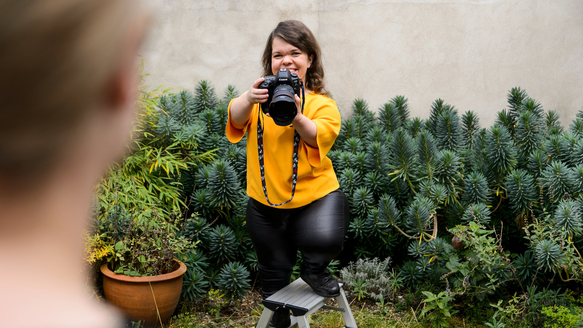 "einfach Mensch: Lasst mich arbeiten!": Anna Spindelndreier während der Arbeit mit Kamera im Garten – im Anschnitt unscharf vorne rechts die Kundin. Anna Spindeldreier steht auf einer kleinen Trittleiter.