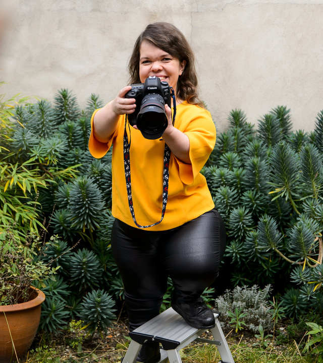 "einfach Mensch: Lasst mich arbeiten!": Anna Spindelndreier während der Arbeit mit Kamera im Garten – im Anschnitt unscharf vorne rechts die Kundin. Anna Spindeldreier steht auf einer kleinen Trittleiter.