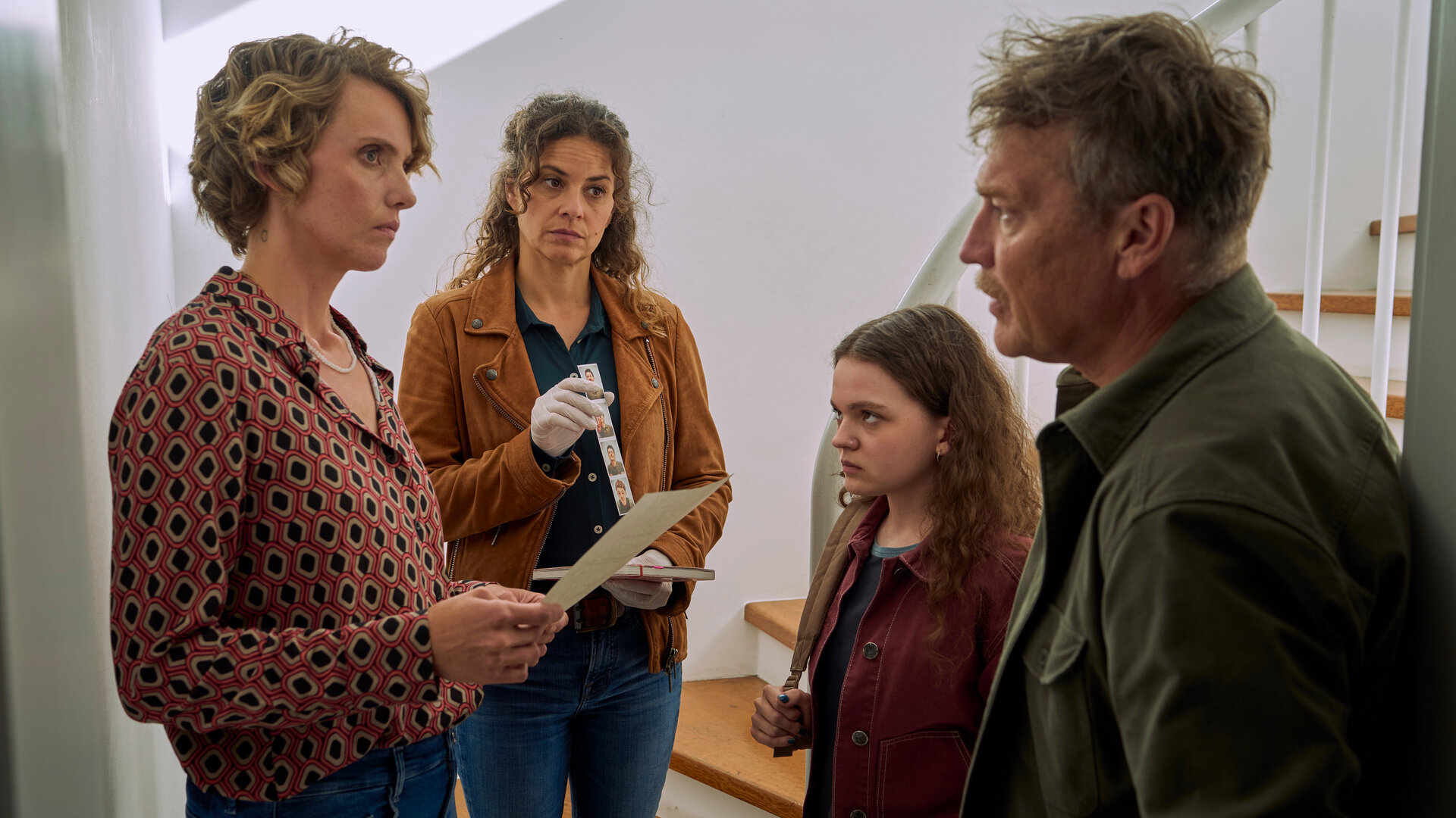 "SOKO Köln - Mobbing mit Todesfolgen": Matti Wagner (Pierre Besson) und Lilly Funke (Tatjana Kästel) stehen in einem Haus vor einer Treppe. Zwischen ihnen stehen sich Katinka Braun (Evelin Schwarz) und Renate Braun (Maike Elena Schmidt) gegenüber. Katinka sieht ihre Mutter grimmig an. Renate hält einen Zettel in den Händen und blickt angespannt zu Matti Wagner. Lilly hält eine Reihe von Passfotos, sowie ein Notizbuch in den Händen. Ihr Gesicht ist nachdenklich.