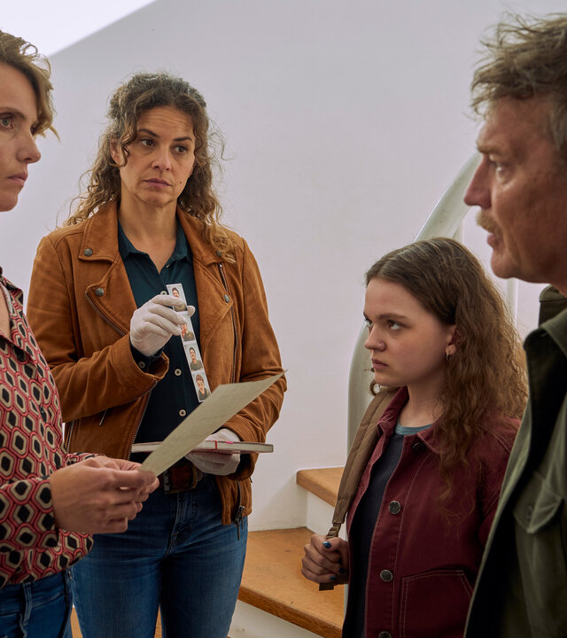 "SOKO Köln - Mobbing mit Todesfolgen": Matti Wagner (Pierre Besson) und Lilly Funke (Tatjana Kästel) stehen in einem Haus vor einer Treppe. Zwischen ihnen stehen sich Katinka Braun (Evelin Schwarz) und Renate Braun (Maike Elena Schmidt) gegenüber. Katinka sieht ihre Mutter grimmig an. Renate hält einen Zettel in den Händen und blickt angespannt zu Matti Wagner. Lilly hält eine Reihe von Passfotos, sowie ein Notizbuch in den Händen. Ihr Gesicht ist nachdenklich.