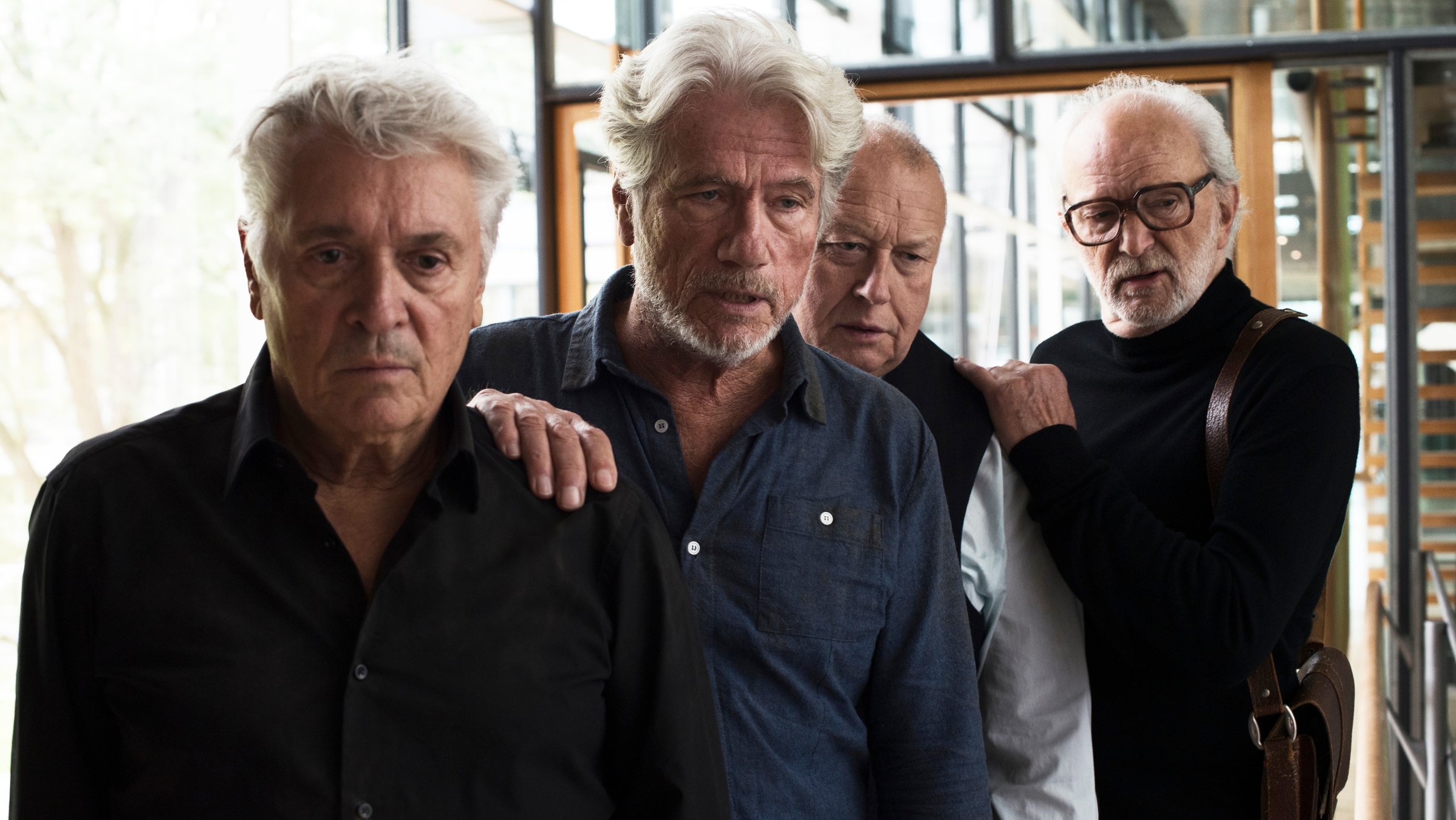 "Kundschafter des Friedens": Jochen Falk (Henry Hübchen), Frank (Jürgen Prochnow), Locke (Thomas Thieme) und Jaecki (Michael Gwisdek) stehen versetzt hintereinander in einem gläsernen Durchgang und schauen erstaunt vor sich.