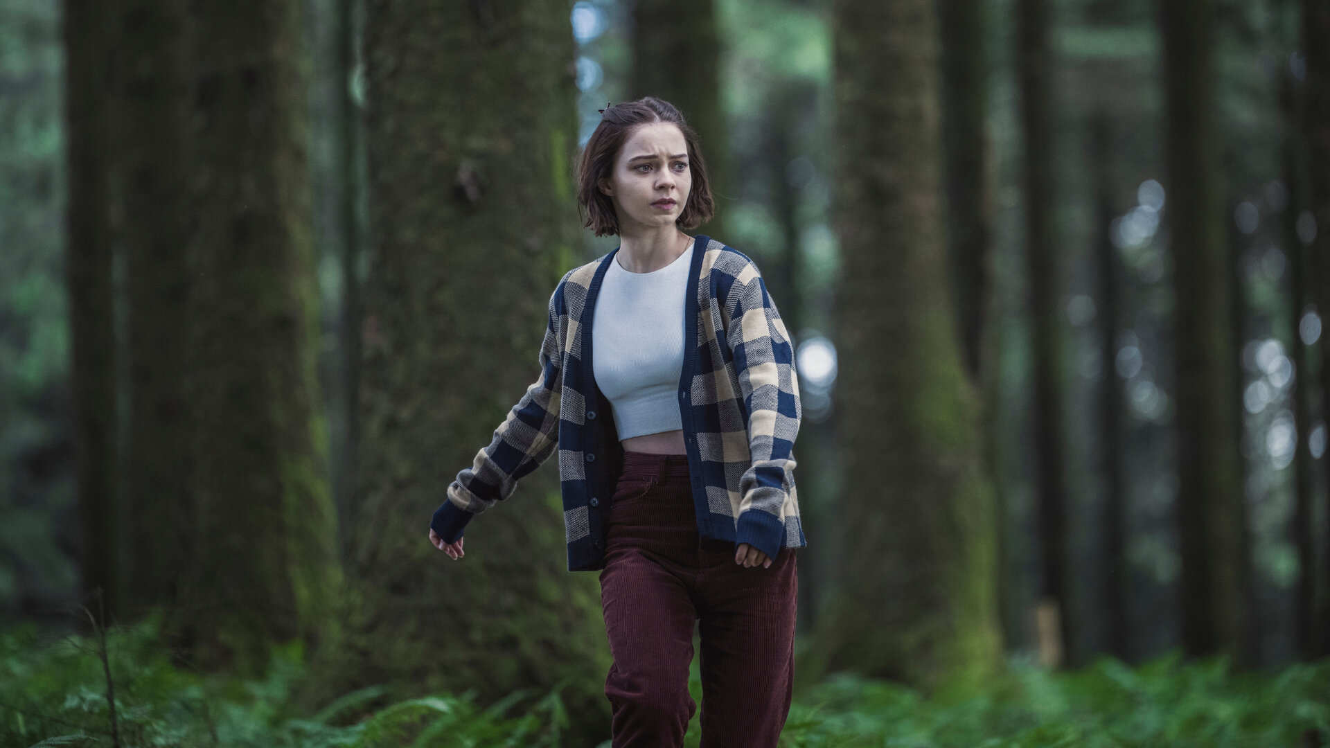 "A Good Girl's Guide to Murder – Eine unglaublich dumme Idee": Pip (Emma Myers) rennt scheinbar orientierungslos durch einen dichten Wald. Sie trägt eine karierte Stoffjacke über einem weißen Top und schaut hilflos, als suche sie jemanden.