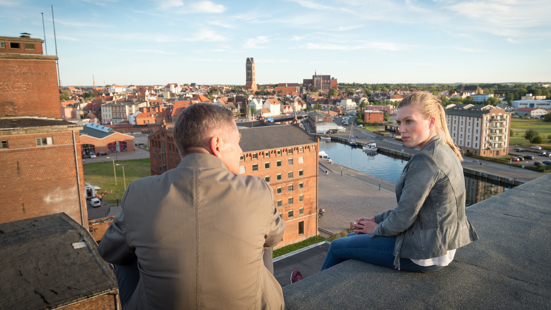 "SOKO Wismar – Das Geständnis": Anneke van der Meer (Isabel Berghout) und Dr. Olaf Bode (Udo Schenk) sitzen mit weitem Blick auf die Hansestadt Wismar auf einem Dach. Sie blicken einander an und unterhalten sich.