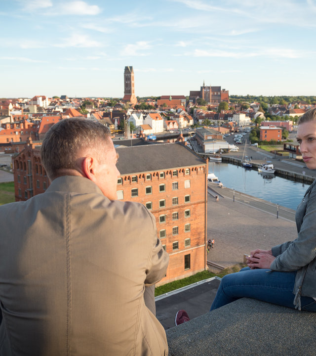 "SOKO Wismar – Das Geständnis": Anneke van der Meer (Isabel Berghout) und Dr. Olaf Bode (Udo Schenk) sitzen mit weitem Blick auf die Hansestadt Wismar auf einem Dach. Sie blicken einander an und unterhalten sich.